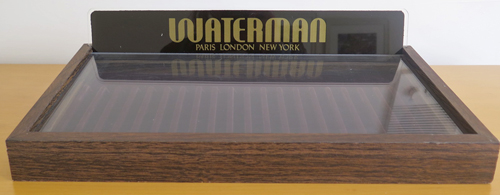ITEM #6388: WATERMAN 20 SLOT DISPLAY CASE WITH PLEXI GLASS TOP. UPRIGHT WATER MAN LOGO. REMOVE THE LOGO PIECE TO REMOVE THE PEN TRAY. SLOTS COVERED IN DARK BROWN VELOUR, SHOWS SLIGHT WARE IN PLACES. "DISPALY UNIT NO. W20"
